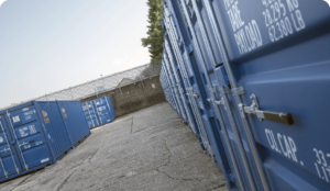 Rows of blue shipping containers aligned on a concrete surface, with one door open, revealing storage space inside. A chain-link fence and some trees are visible in the background under a clear sky.
