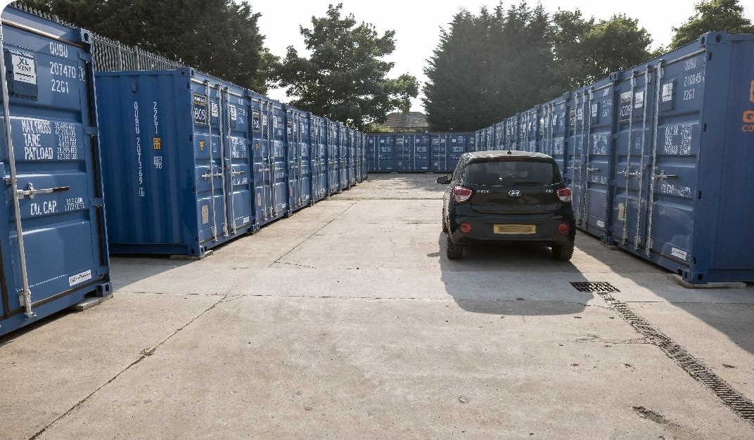 A small black car parked in an outdoor storage facility, surrounded by rows of blue shipping containers on both sides. Trees are visible in the background under a partly cloudy sky.