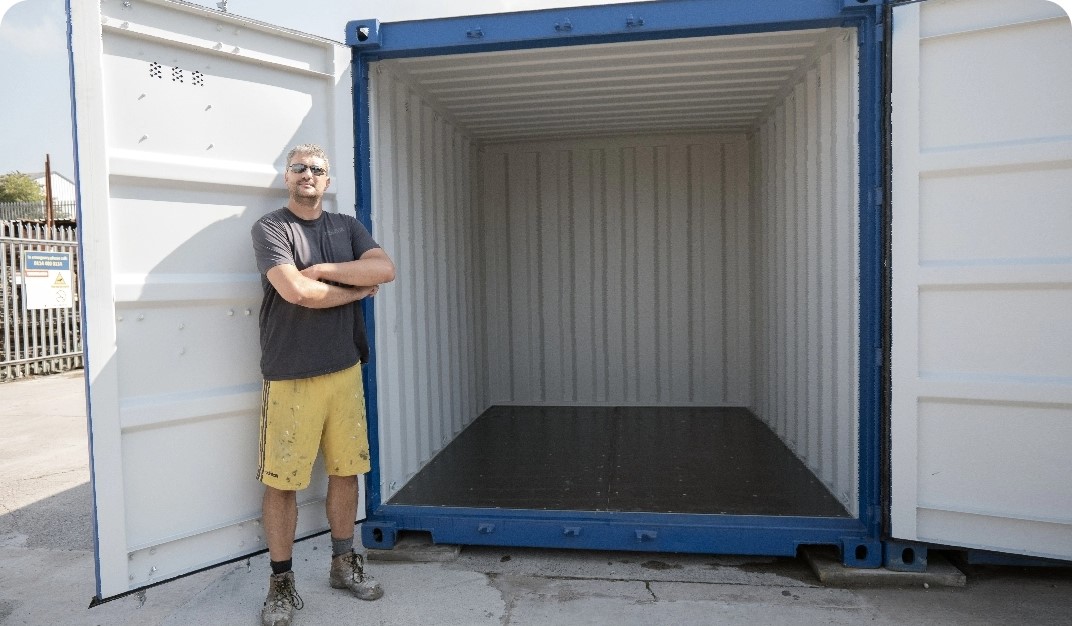 A person with short hair and sunglasses stands with folded arms next to an open, empty shipping container. The container is white with a blue door and is situated outdoors on a sunny day, near a chain-link fence and industrial area.