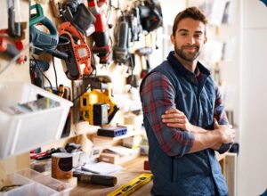 A man with a beard and checkered shirt, wearing a blue vest, stands confidently with arms crossed in a workshop. Various tools and equipment are organized on the wall behind him. A spirit level and a paint can are on the workbench.