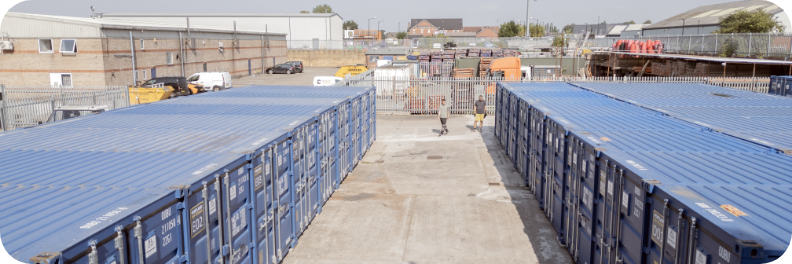 A storage facility named Portland Self Storage features rows of large blue shipping containers. The sky is clear, and the area includes buildings and a few vehicles. A person walks a dog in the open space between the containers.