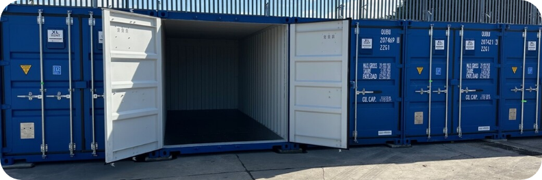 Several blue shipping containers are lined up on a concrete surface at Portland Self Storage. One container is open, revealing an empty interior. White doors contrast with the blue exteriors, while a corrugated metal fence looms in the background.