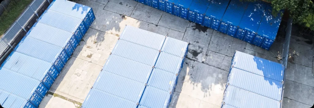 An aerial view of Portland Self Storage reveals a storage yard with rows of neatly arranged blue shipping containers on a concrete surface. Shadows cast by nearby trees and fencing add depth to the scene.