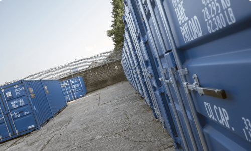 Rows of blue shipping containers aligned on a concrete surface, with one door open, revealing storage space inside. A chain-link fence and some trees are visible in the background under a clear sky.