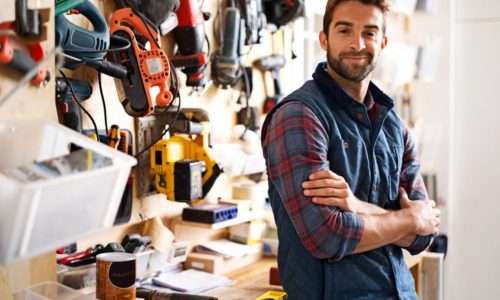 A man with a beard and checkered shirt, wearing a blue vest, stands confidently with arms crossed in a workshop. Various tools and equipment are organized on the wall behind him. A spirit level and a paint can are on the workbench.
