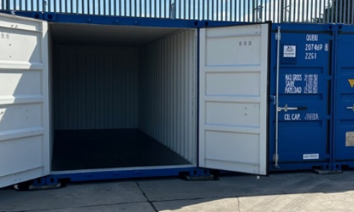 Several blue shipping containers are lined up on a concrete surface at Portland Self Storage in Sheffield. One container is open, revealing an empty interior. White doors contrast with the blue exteriors, while a corrugated metal fence looms in the background.