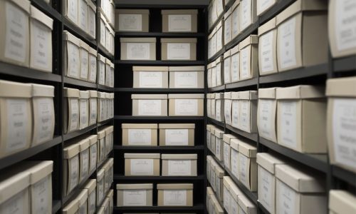 A narrow archive room filled with rows of beige document storage boxes neatly arranged on black metal shelves, labeled and organized for record-keeping.