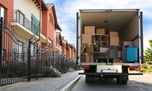 A moving truck is parked on a residential street with its back open, revealing various packed items like boxes, a chair, a rolled carpet, and a blue suitcase. Rows of brick houses and an iron fence are visible in the background.