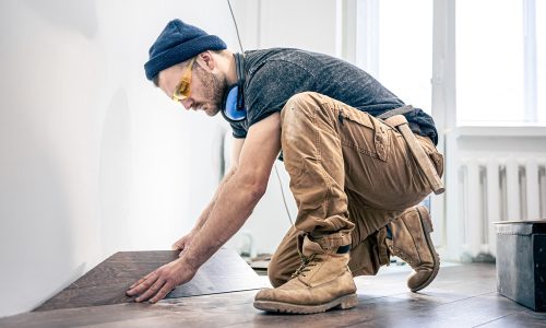 A person in work boots, cargo pants, and a beanie is kneeling while installing wooden flooring. They are wearing protective eyewear and ear protection, focused on aligning a floorboard. Sunlight streams through a window in the background.