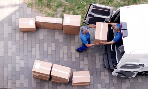 Two people wearing blue uniforms unload cardboard boxes from a white van onto a paved walkway. The boxes are arranged in a curved line, and there is grass visible in the background. The scene is viewed from above.
