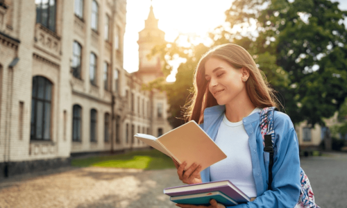 A young woman stands outside a university building, smiling as she reads from a notebook. With textbooks in tow and her blue shirt peeking from beneath a hefty backpack, she embodies the promise of new beginnings. The sunset casts a warm glow, capturing a moment worth storing in memory.
